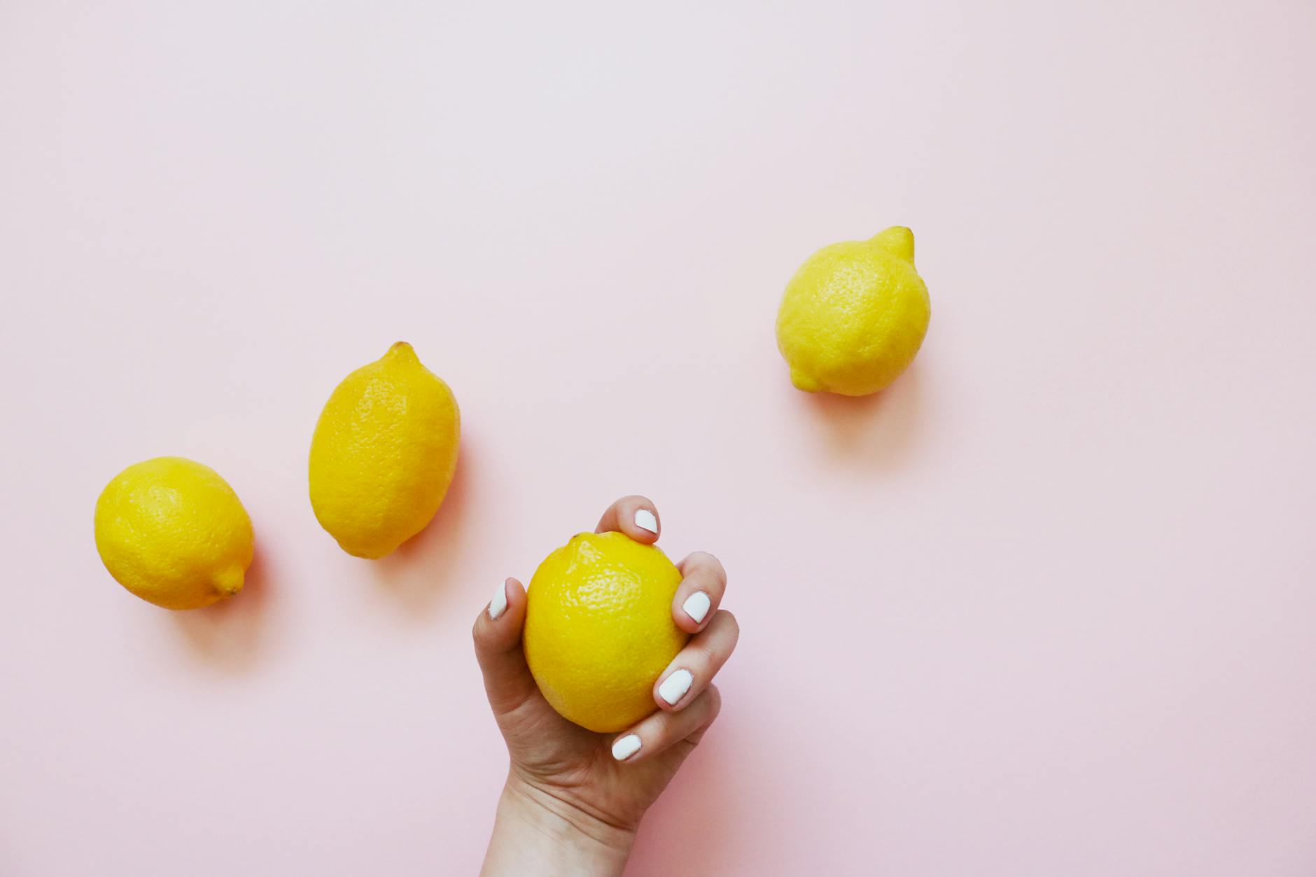 A hand holding a fresh lemon against a soft pink background